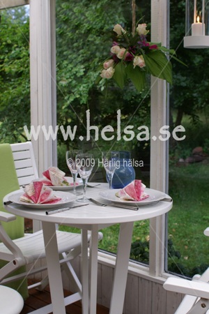 En sommardukning på verandan för tre personer. Vitt porslin, rosavita servetter och vitvinsglas. Över bordet hänger en blomsteruppsättning med bland annat vita rosor.
